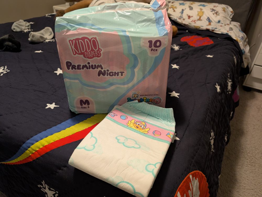 A bag of opened Kiddo Premium Night diapers sitting on my bed, with one diaper lying in front of it. It's a cloth-backed diaper with the cloud outlines on it and a narrow pink tape strip with a happy-looking yellow bird character in the middle.