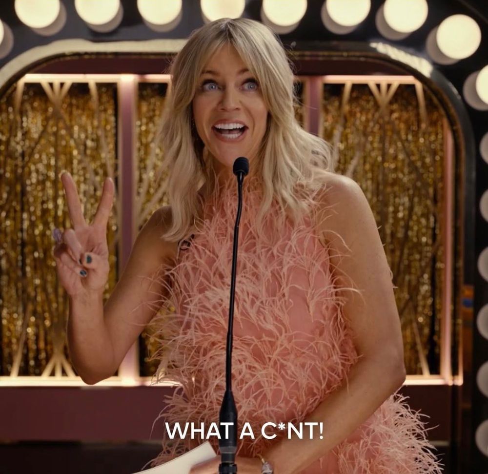 Screenshot from tv show Hacks where actress Kaitlin Olsen as DJ is on stage at the roast of her mom. She’s giving a peace sign with a smile on her face and is saying, “What a Cunt!”