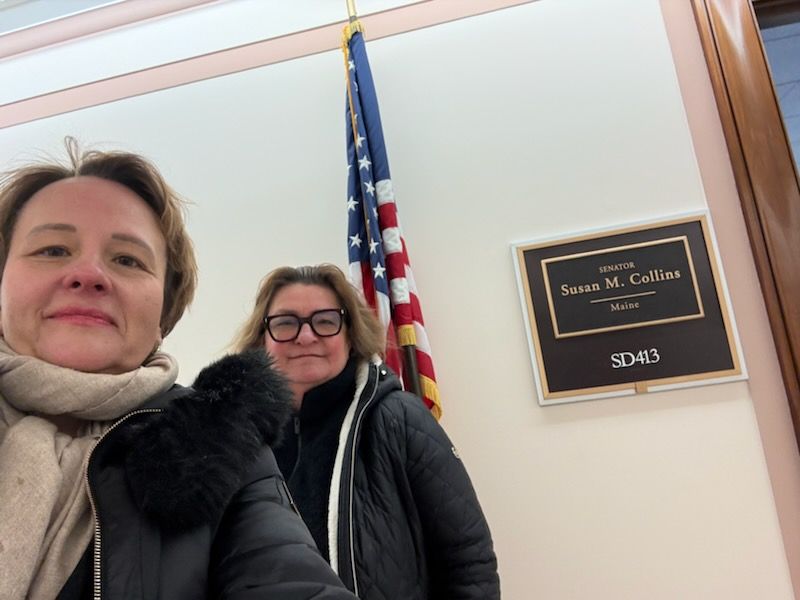 Two scholars delivering a letter to Senator Susan Collins' office, American flag in background, official seal visible