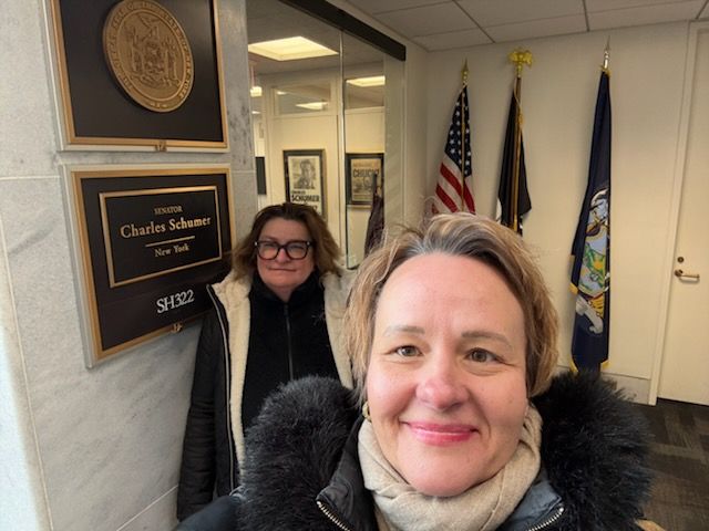 Two scholars delivering a letter to Senator Chuck Schumer's office, American flag in background, official seal visible