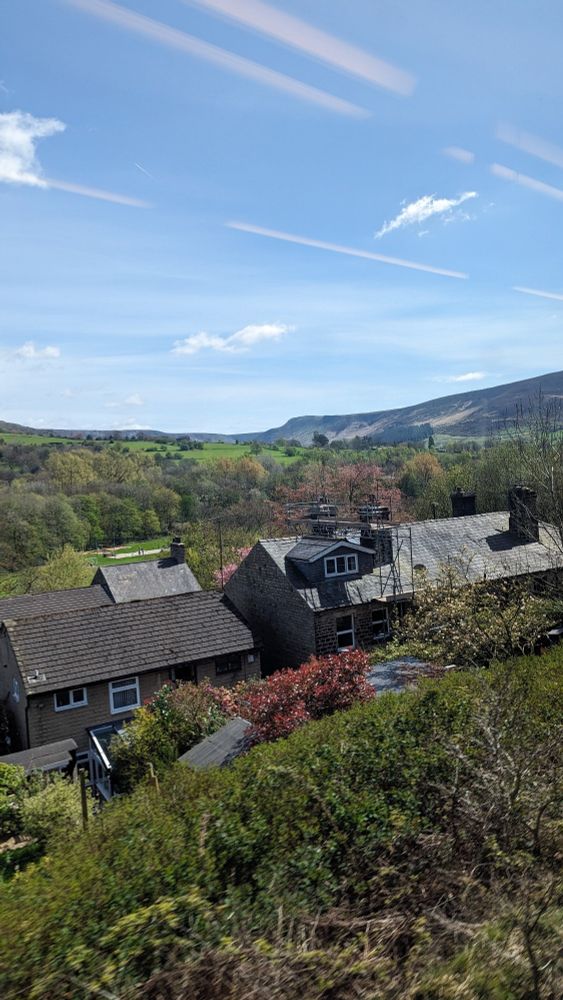 Photograph of English countryside, clear, sunny blue skies with hills framing the background. There are green fields toward the hills, then trees, and then some brick houses close to the camera that have construction on the roof.