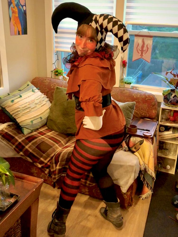 A photo of myself in a jester costume with patterns and lots of ruffles. The background is a room with large windows, a couch, and blankets.
