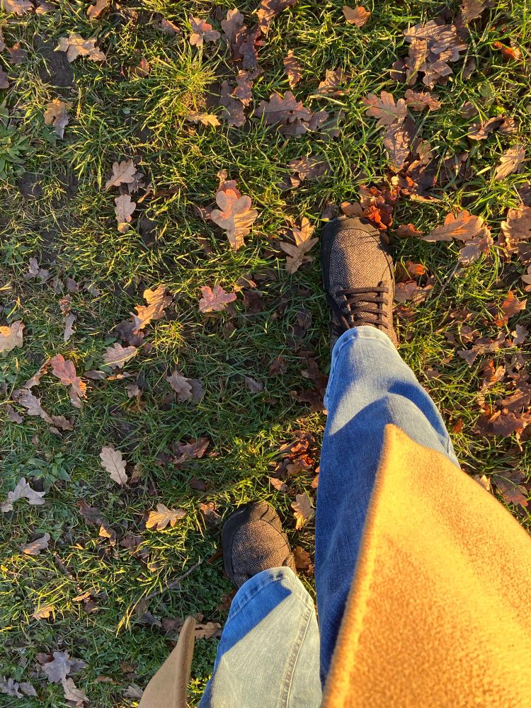 Blick auf meine Beine in Jeans und braunen Schuhen auf Gras, über dem rechten Bein Teil eines gelben Mantels