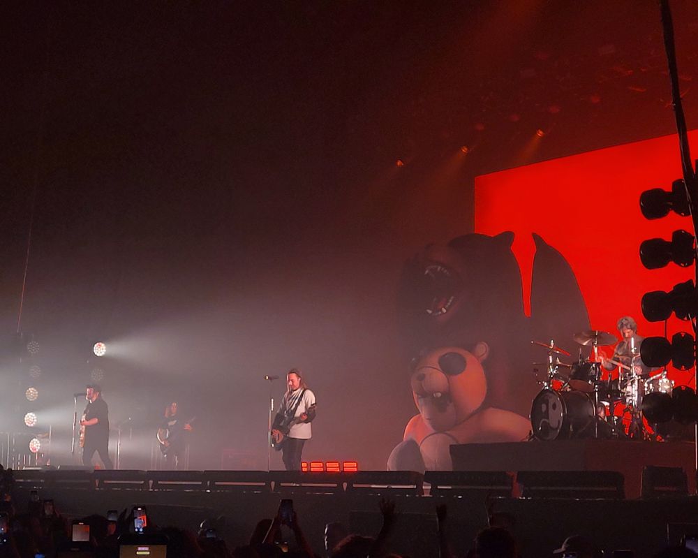 foto do show do fall out boy. no palco, dá esquerda para direita, temos o patrick tocando guitarra, o pete tocando baixo e o andy tocando bateria. ao fundo, um boneco inflável de um garoto vestido de urso carregando um urso nas costas, em referência a capa do álbum folie à deux. as luzes do palco estão vermelhas.