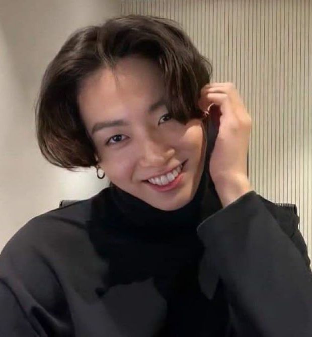 jungkook se sentindo formidável com a mão na orelha, rindo pra câmera e usando uma blusa de manga comprida preta