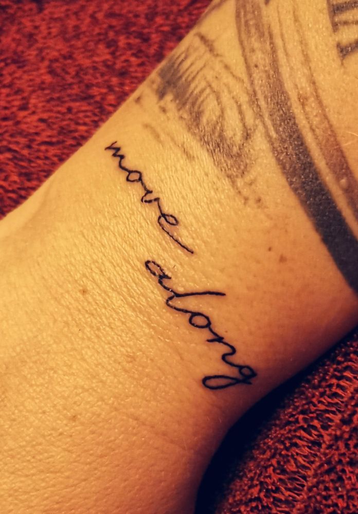 Der Schriftzug 'move along' auf einem Handgelenk tätowiert. Der Arm liegt auf einer roten Decke. Über dem Schriftzug ist ein Teil eines weiteres Tattoos zu sehen.