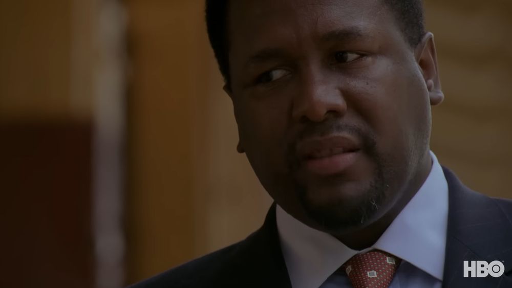 A Black man, actor Wendell Pierce, casts a disgusting look off-camera. He is wearing a suit with a blue shirt and a red tie. The screenshot is from the HBO series "The Wire."