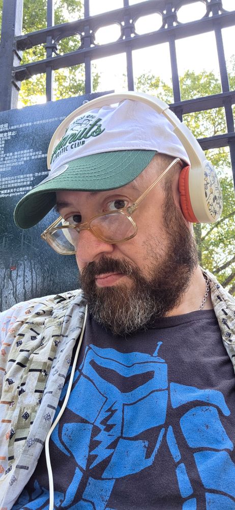 Your skeeter, a bearded, bespectacled, cis "White" man, sits outside wearing a cap & headphones emblazoned with Snoopy, the iconic beagle from Charles M. Schulz's landmark comic strip PEANUTS, as well as a charcoal grey tee ft a blue robot.