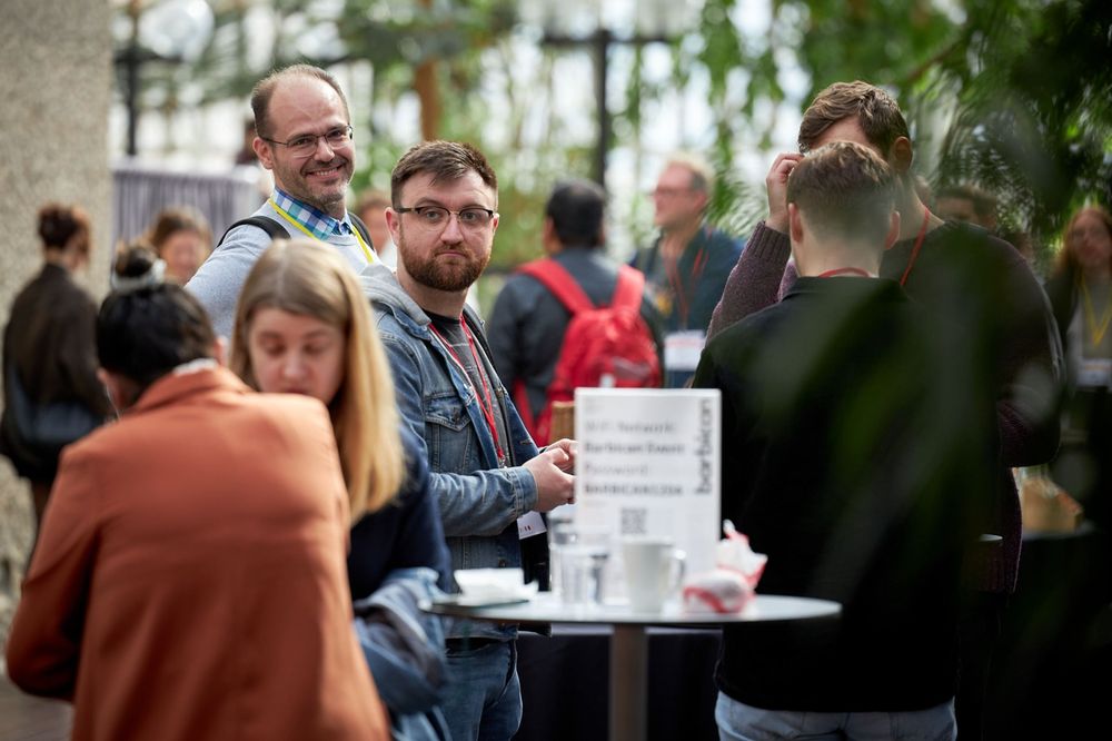 A group of State of the Browser attendees at a break chatting around standing-height round tables in the conservatory area of the Barbican Centre in London. In focus is a group of four men, one of which is me looking directly at the camera smiling because I spotted the photographer, Marc Thiele, trying to take candid photos of the crowd.
