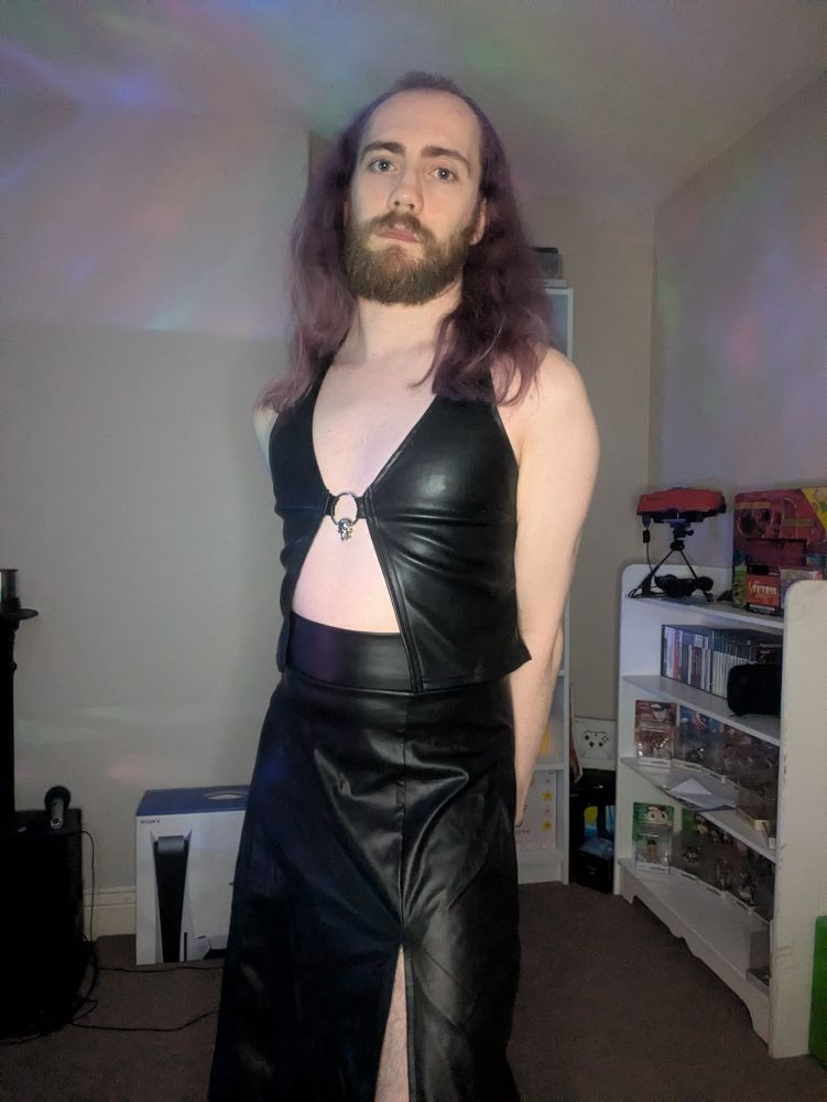 Blank Master wearing a faux leather halter top and faux leather skirt, with their hands behind their back. Multi-coloured caustics are being projected onto the scene.