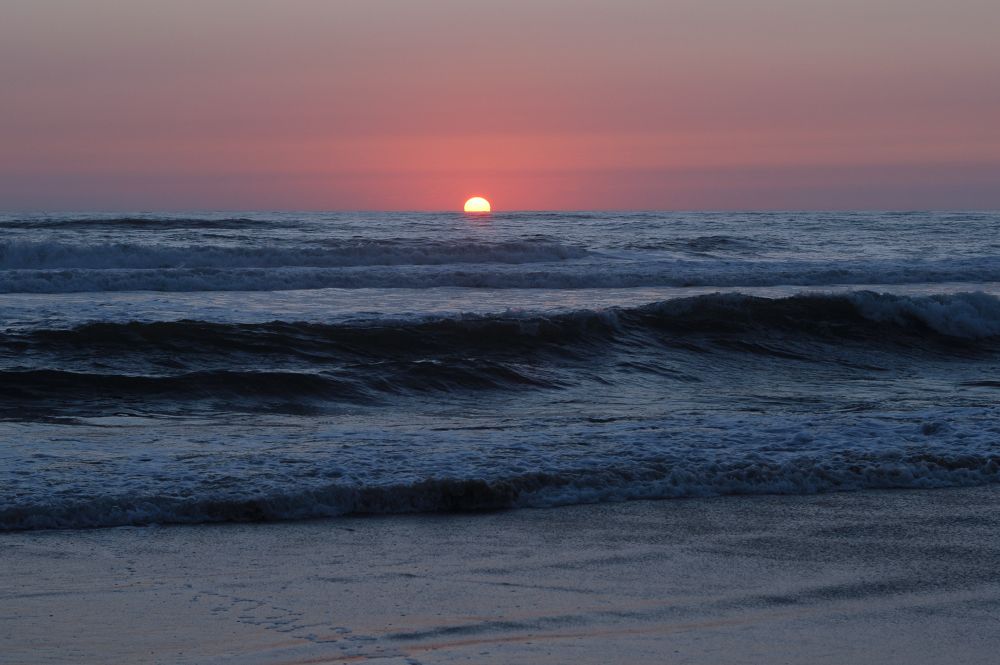 A picture of the bright orange Sun just little over halfway above horizon, in a sky of salmon gradating to pink hues with darker colored overcast at the margins. The middle ground shows the Atlantic Ocean with gray waves crashing on the sandy shore in the foreground. 