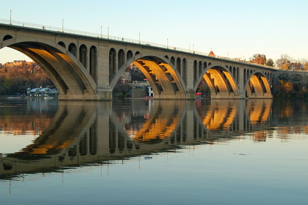 A picture of 4 arches of Key Bridge ending on the DC side of the Potomac, in the good light of late afternoon Sun. On a calm day with the water reflecting the bridge and a clear light blue sky.      