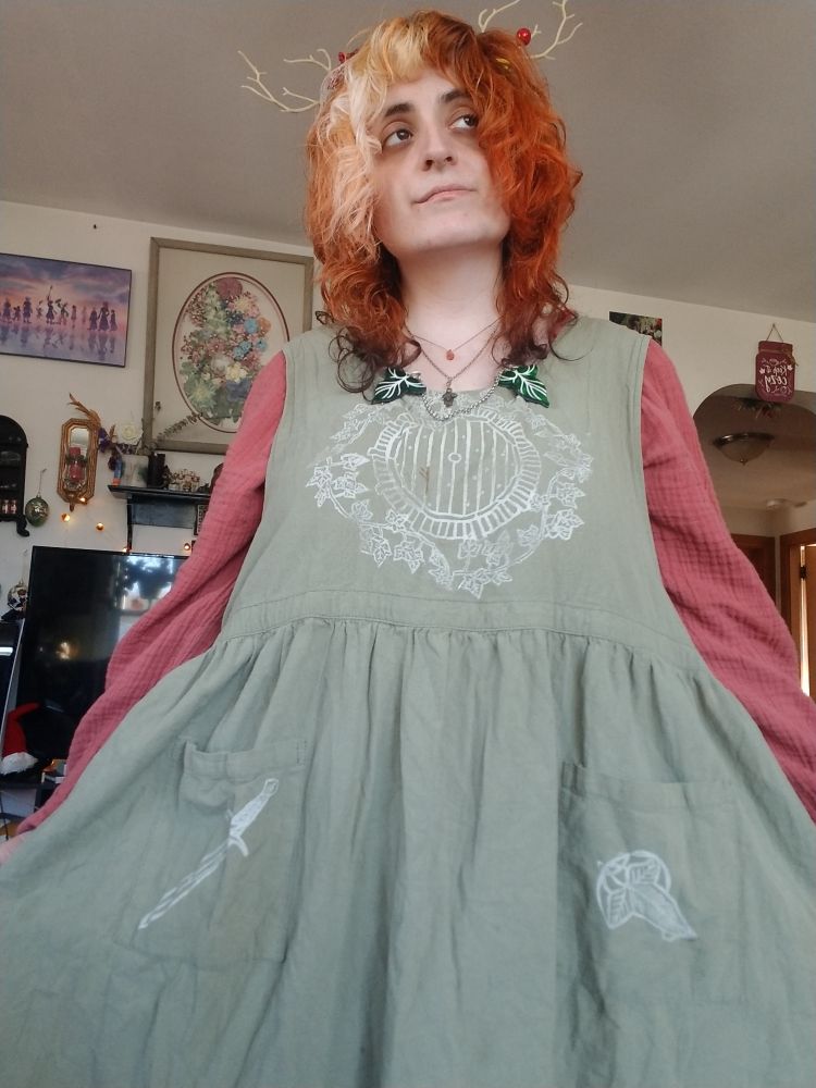 Fern stands holding out her sage green apron. It has white stamped details from lord of the rings, the chest being a hobbit door. Under it she is wearing an orange long sleeved dress. Clipped to the front of her apron is two acrylic lord of the rings leaves with a silver chain between. For jewelry she is wearing a sunstone drop and a metal oak leaf with acorns