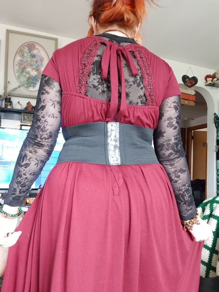 Fern from the back, showing a rectangle cutout in the back of her wine red dress over her shoulders. There is a tie tied in a bow. She has on a black bodysuit with black velvet flowers. She also has on a black faux corset belt