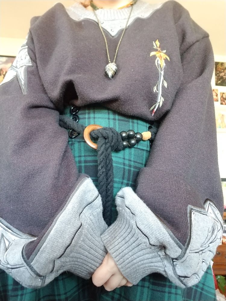 A close up of Fern clasping her hands infront of her to show off her sweater, which has embroidery that looks like armor, and a sword embroidered on the chest. 