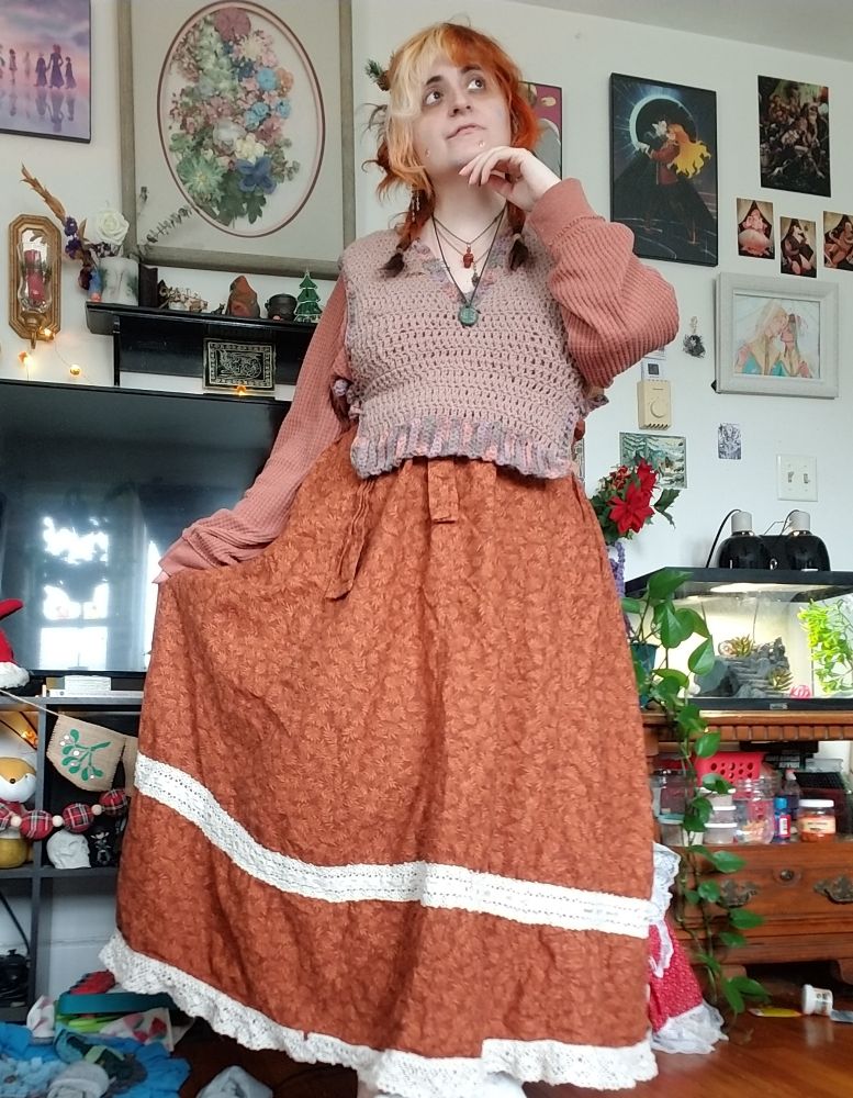 Fern stands with one hand to her chin, the other holding out the burnt orange wrap skirt she is wearing. It has cream lace trim. On top she has on a taupe crocheted tie side vest over a slightly less saturated burnt orange waffle shirt. 