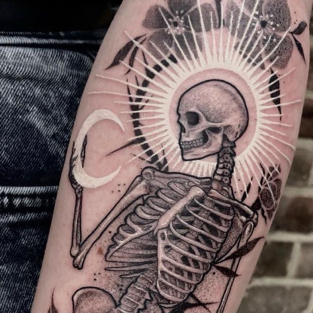 A black and white ink tattoo of a skeleton holding a crescent moon, with a halo and a curved branch of cherry blossoms surrounding its head.