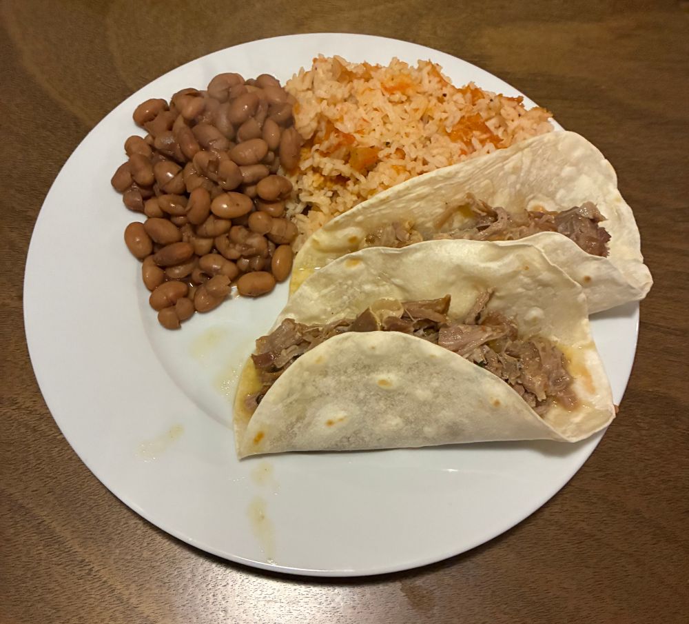 a white plate with two carnitas tacos, rice, and beans