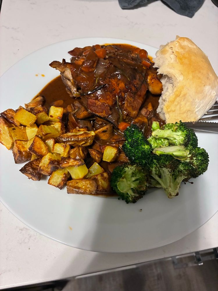 a picture of coq au vin on a plate. a chicken thigh is covered in a thick brown red wine sauce with carrots and veggies. on the plate is also roasted potatoes and roasted broccoli and some sad looking homemade bread