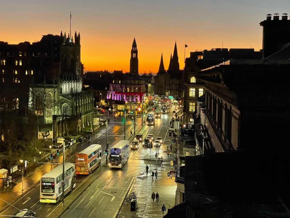 Edinburgh skyline at west end Princes Street, silhouetted against orange glowing sky, spire of Charlotte Chapel is central, wet streets and traffic in foreground.