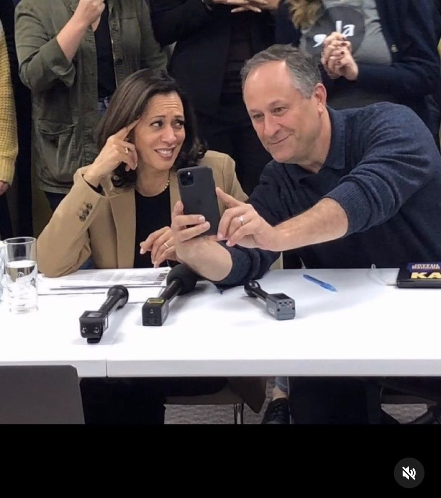 Doug showing Kamala something on his phone; he looks pleased, while she is giving him a disgusted sidelong look