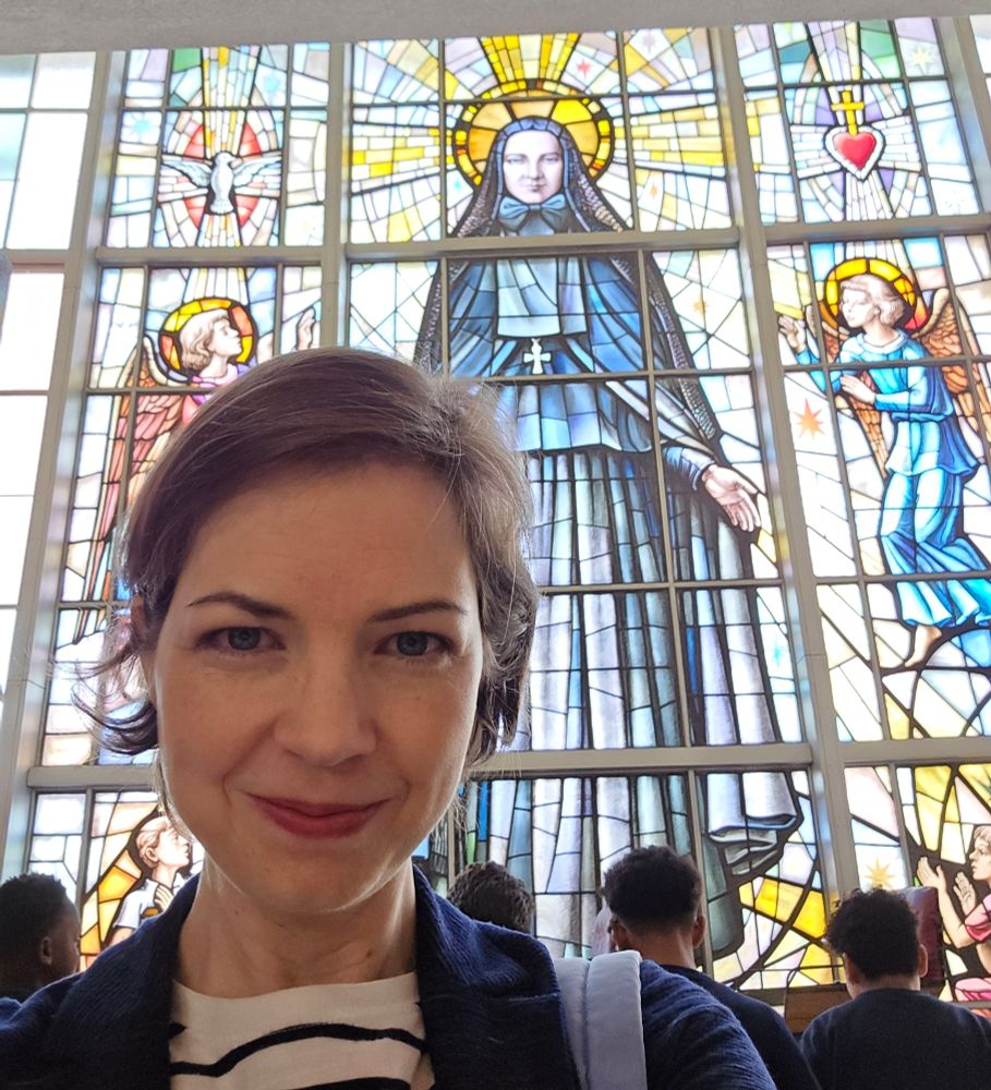 Me in the foreground, in front of a very large stained-glass window depicting St. Frances Xavier Cabrini, patron of immigrants, in glory, as they say