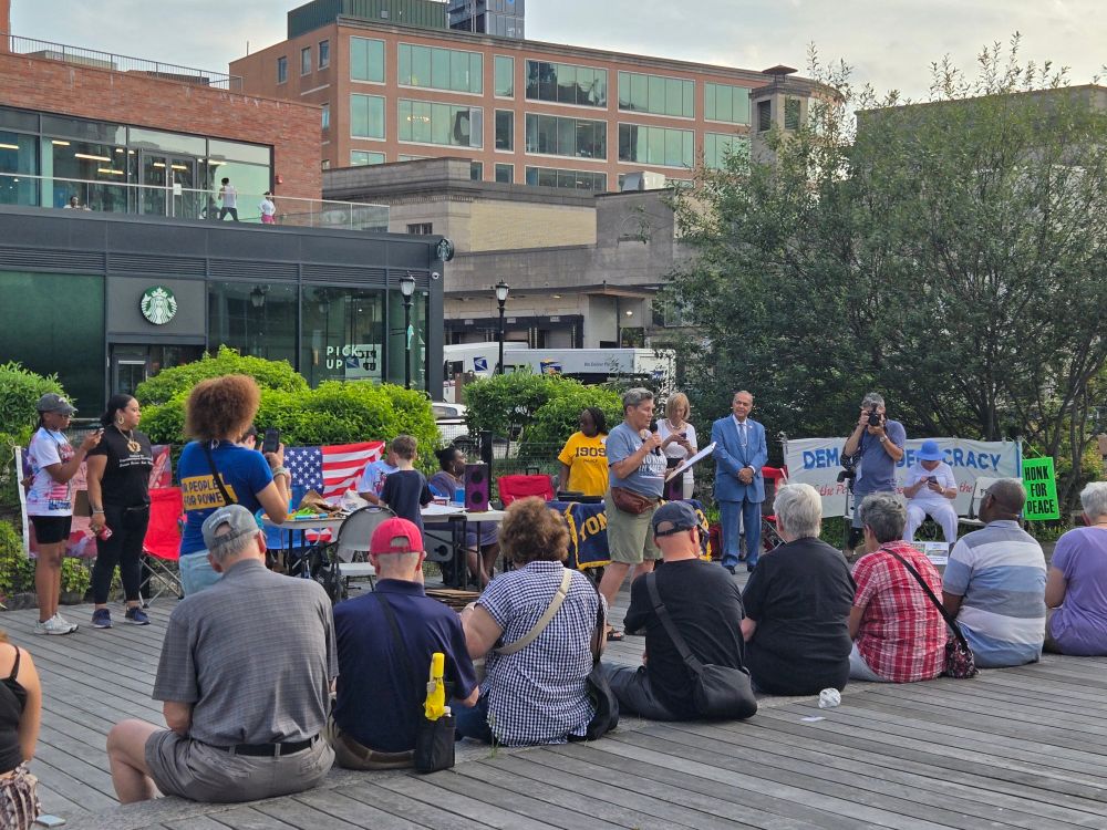 Some people assembled in Van Der Donck Park in Yonkers, listening to a rally organizer speaking into a microphone. Flags, signs, politicians, and a Starbucks in the background.