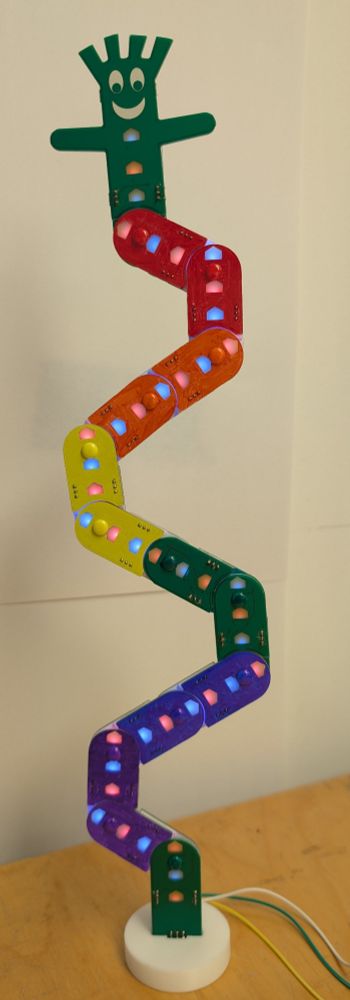 A tabletop display of a multi-segmented car wash balloon man sculpture constructed of PCBs in different colors arranged at jaunty angles to each other with some the backlit LEDs running up the front center of the sculpture illuminated in an alternating blue and red pattern.