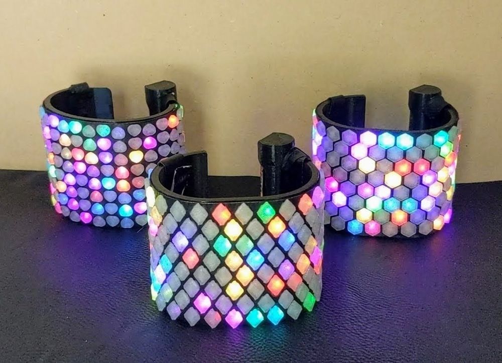 Three Illuminated cuff bracelets with bright colorful patterns illuminating 3D printed grids comprised of geometric shapes on the exterior. One grid is made of heart shapes, one of diamond shapes, and one of hexagons.