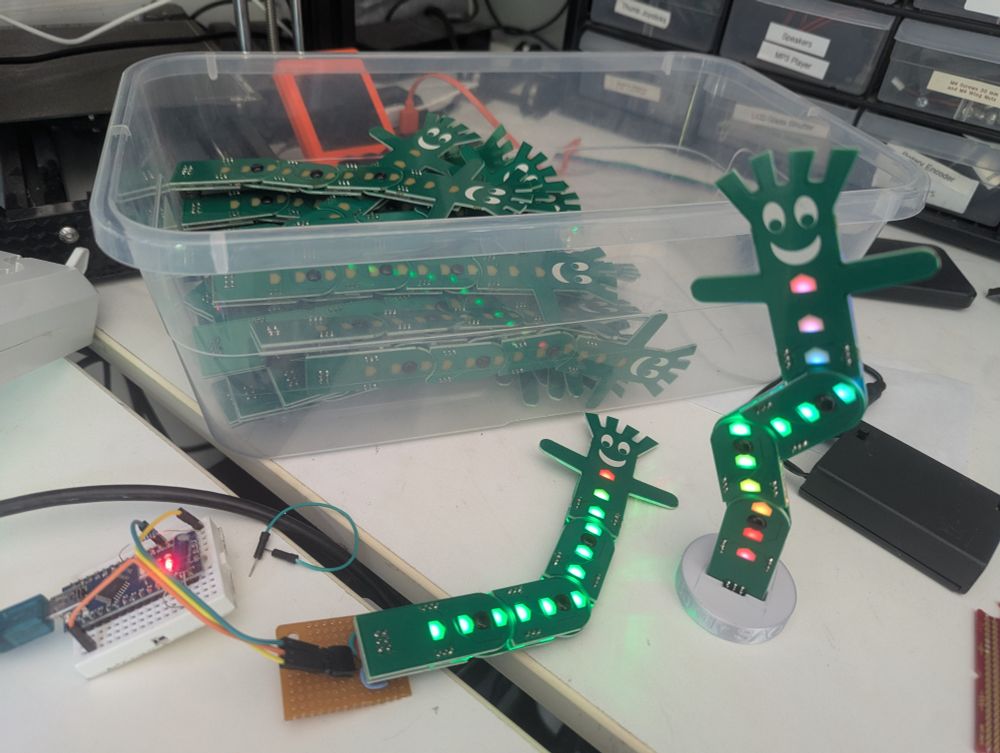 Two green poseable PCB assemblies, shaped like inflatable car wash men are illuminated through translucent patches on the front of the PCBs. One assembly is laying on a table, connected to a programmer while the other assembly stands vertically with its base held by a round silver 3D printed stand. Begind the assemblies is a plastic bin filled with dozens more identical assemblies.