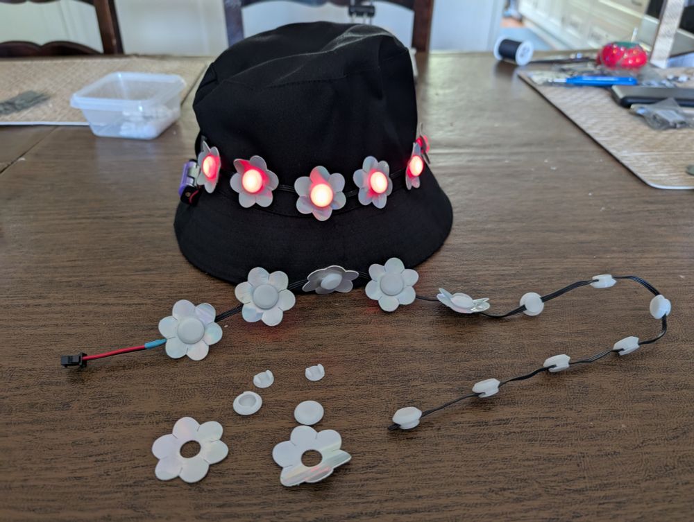 A black bucket cap rests on a table. Several silver flowers with illuminated red centers run around the middle of the hat. In front of the hat is a string of pebble lights in which the LEDs have been covered by translucent circular diffusers. Some of the diffusers form the center for vinyl flowers that have been placed around them. On the table in front of the LED string are two of the 3D printed diffusers, along with two flowers cut from vinyl with holes in their centers for placing the diffusers through.