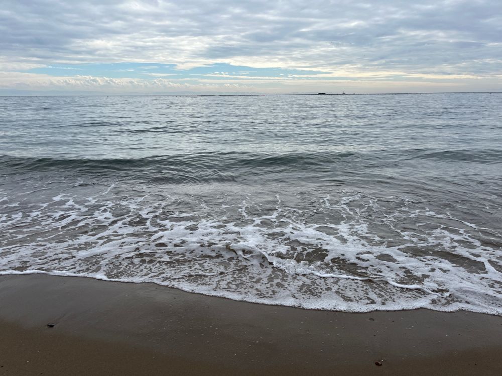 昼間の海岸で白波が砂浜に押し寄せている。空はほぼ曇りだがところどころ青空がある