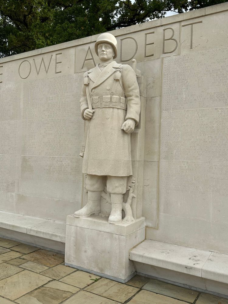 A statue of a soldier in helmet with grenades and ammunition pouches at Madingley American cemetery . Owe A Debt written on the wall behind 
