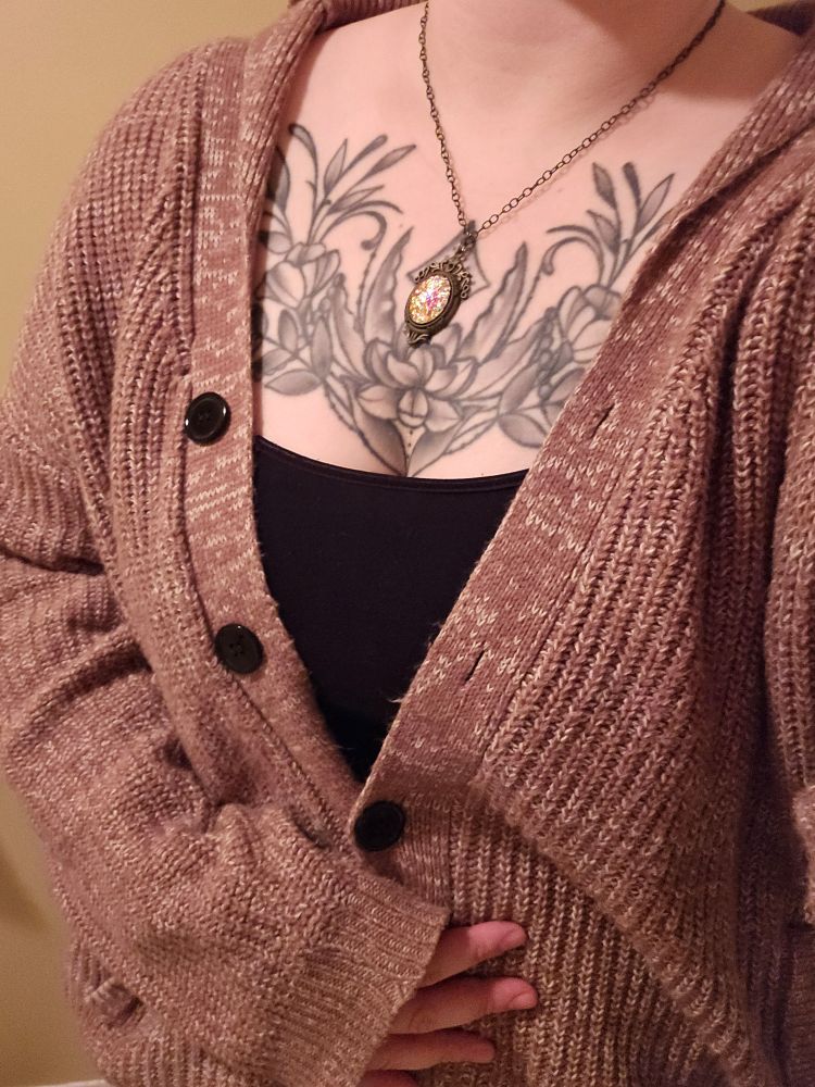 A faceless selfie of a person wearing a black tanktop and a large brown cardigan with a plant tattoo on their chest and an open pendant necklace. 