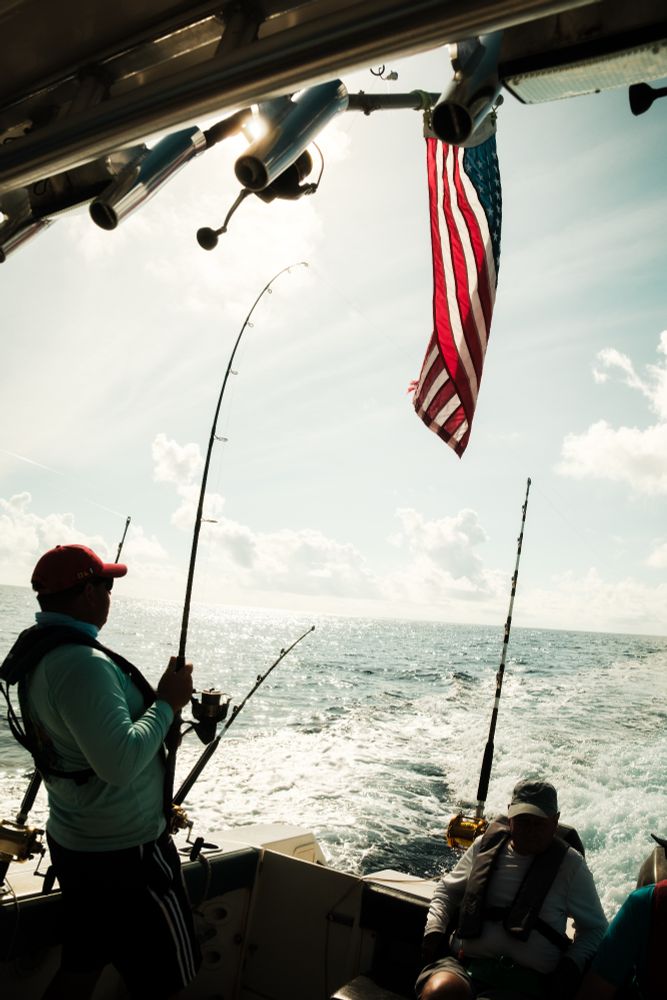 Anglers aboard a boat fish in the open waters of the British Indian Ocean Territory. One person stands reeling in a catch with a curved fishing rod, while others are seated nearby. The U.S. flag flies above the stern as the boat moves through calm, sunlit seas under a partly cloudy sky.