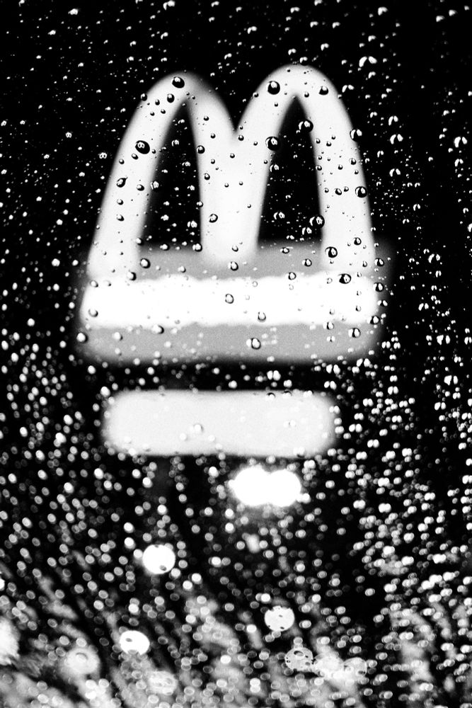 Title: ғᴀsᴛ ғᴏᴏᴅ ʀᴇғʟᴇᴄᴛɪᴏɴs – ʀᴀɪɴʏ ᴅʀɪᴠᴇ-ᴛʜʀᴜ ɴɪɢʜᴛs 

Raindrops on a windshield blur a glowing fast food sign in the background, creating an abstract black and white night scene.

The glow of comfort food through a windshield of rain—nothing says American midnight like fast food lights and the hush of a rainy drive-thru lane.

Black and white photo capturing the abstract beauty of a fast food logo (suggestive of McDonald’s golden arches) seen through a rain-covered car window. The image evokes themes of nostalgia, late-night cravings, American roadside culture, and rainy urban drives. Ideal for fans of street photography, fast food aesthetics, car culture, and rainy reflections.