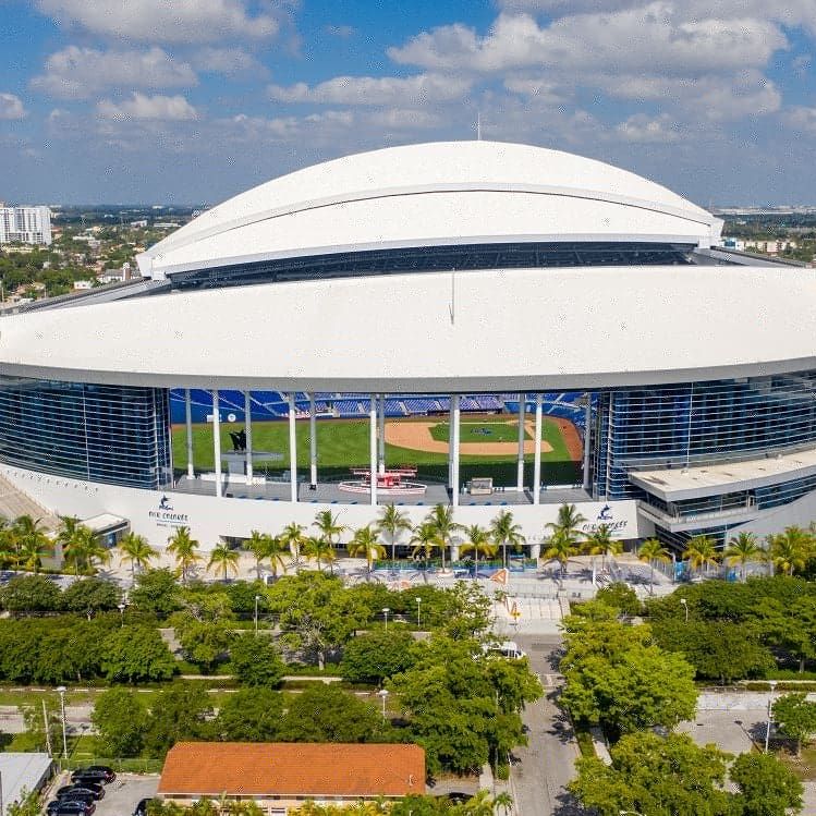Photo 2 (Baseball stadium):
Aerial view of loanDepot park in Miami, Florida, home of the Miami Marlins, featuring its closed retractable roof, palm tree-lined entrance, and visible baseball field under sunny skies.
