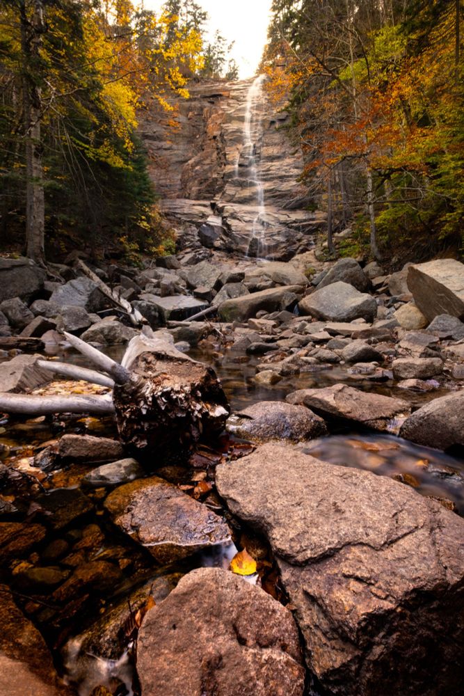 Waterfall and Forest: Scenic autumn waterfall cascading down a rocky cliff surrounded by colorful fall foliage in a forest in New England. Clear stream with boulders and fallen logs in the foreground.