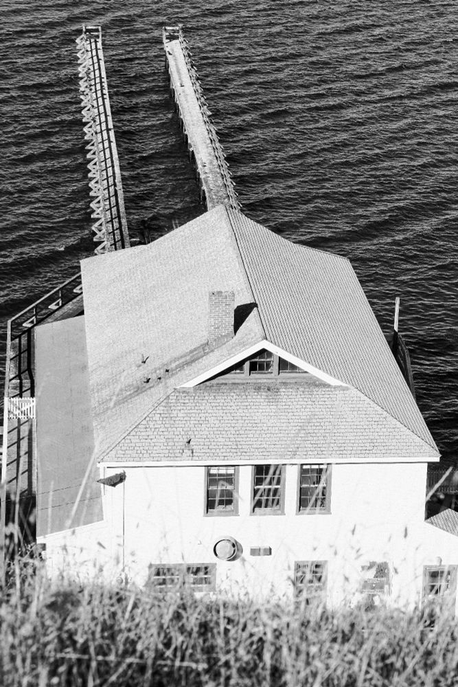 Black-and-white photo looking down toward a historic coastal structure with a steep roof extending toward two long, narrow wooden piers reaching into dark, rippling ocean water. The perspective captures the geometry of the roof and symmetry of the piers, evoking a timeless maritime feel. (Keywords: coastal architecture, pier, ocean texture, black and white photography, maritime history.)