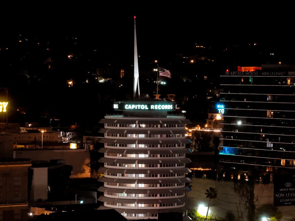 Night view of the iconic Capitol Records Building in Hollywood, California, illuminated with "CAPITOL RECORDS" signage at the top. The cylindrical, stack-like architecture stands out against the dark hillside backdrop, with an American flag flying above and surrounding city lights visible in the distance.