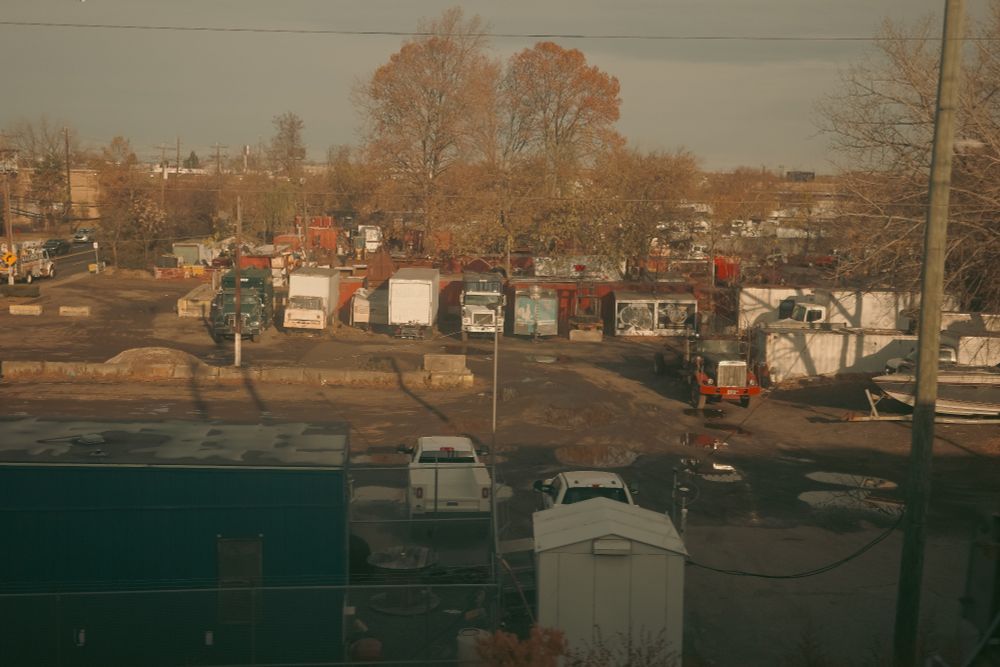 Industrial yard with trucks and equipment, surrounded by leafless trees under soft golden-hour light, viewed through a train window.