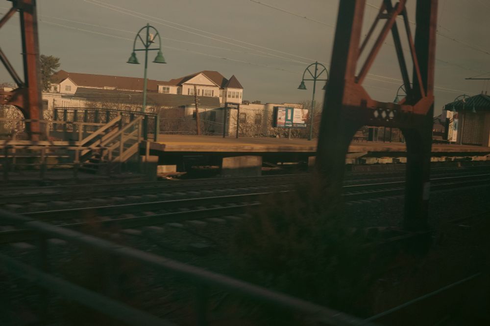 Small-town train station platform, seen from a moving train, with vintage-style green lamp posts and residential buildings in the background.