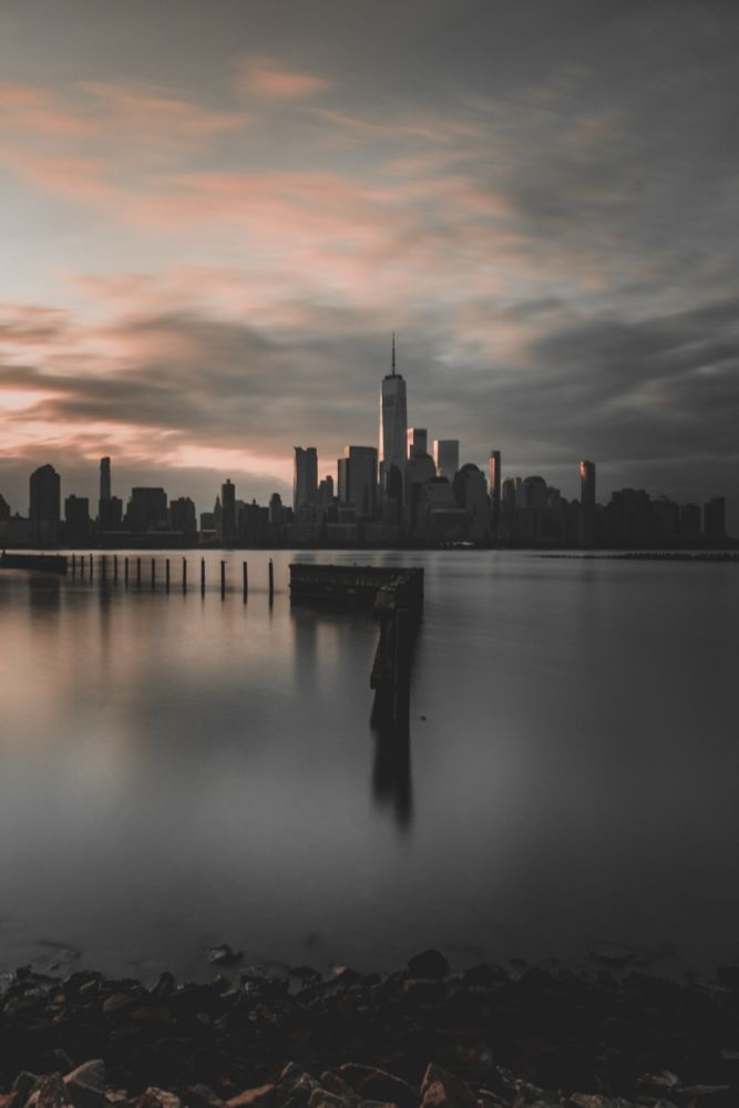 This is a photograph of a city skyline, likely taken around sunrise or sunset, with a body of water in the foreground. The tallest building in the center appears to be One World Trade Center in New York City. The image has a moody and dramatic atmosphere, with soft lighting and long exposure creating a smooth water effect. The warm hues in the sky contrast with the dark silhouettes of the buildings. Let me know if you need any modifications or analysis of the image.