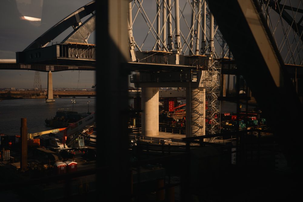 Bridge construction site over a river, captured through a train window, with cranes, scaffolding, and heavy machinery bathed in warm afternoon light.