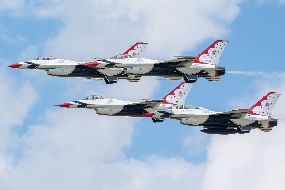 Four U.S. Air Force Thunderbirds F-16 jets fly in tight diamond formation against a bright blue sky with scattered clouds, showcasing precision and power during an airshow performance.