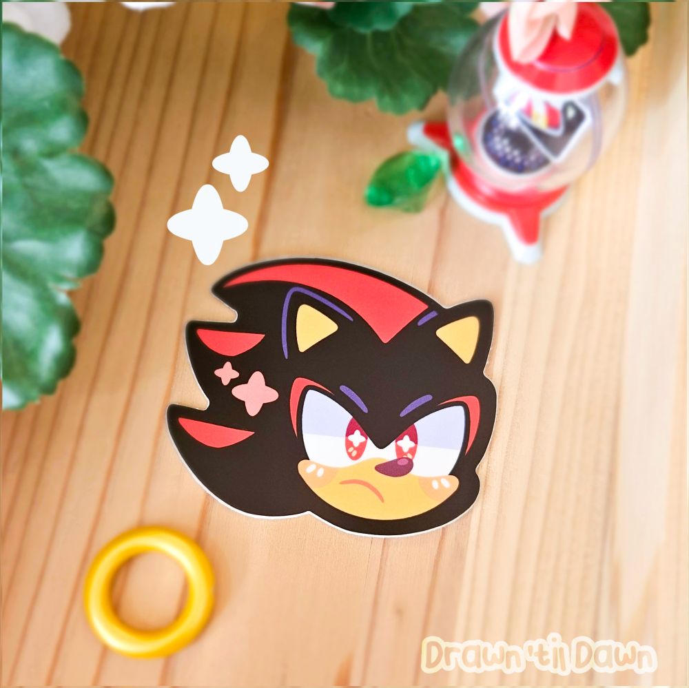 A photo of a Shadow vinyl decal resting on a wooden surface surrounded by green leaves, a ring, a green emerald. and an item bubble from Sonic Adventure.