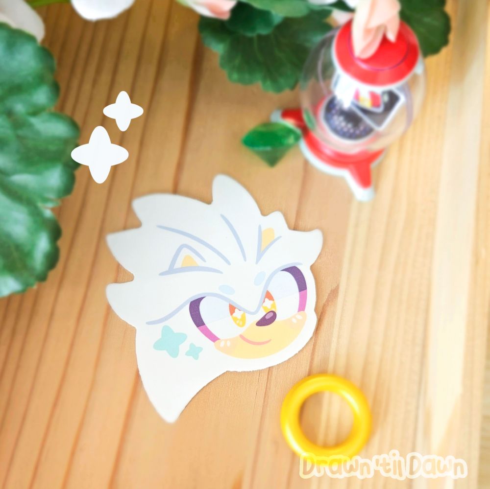 A photo of a Silver vinyl decal resting on a wooden surface surrounded by green leaves, a ring, a green emerald. and an item bubble from Sonic Adventure.