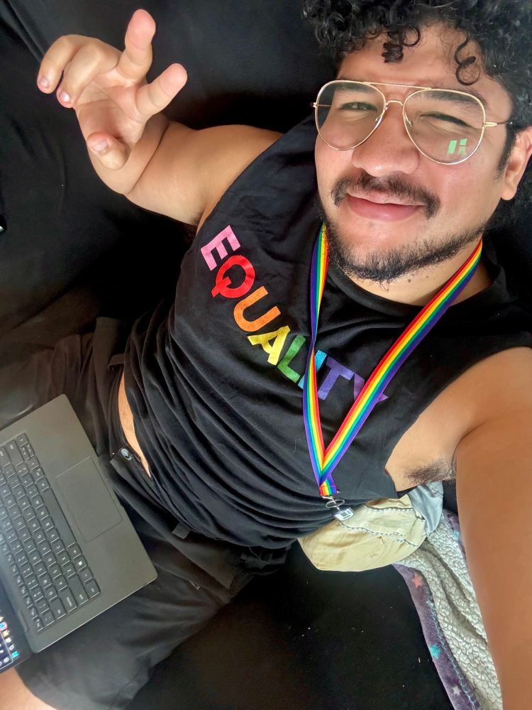 Seated selfie with laptop open on lap wearing black tanktop with Equality written on it in rainbow colors