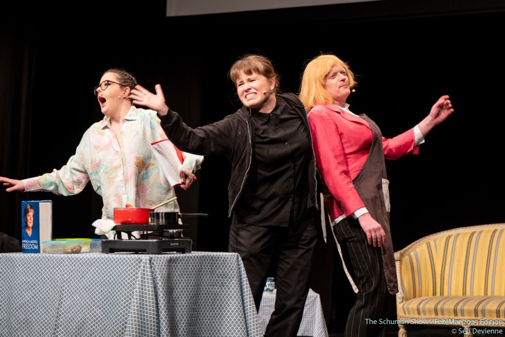 Three cast members on stage, with one dressed up as Angela Merkel, being swept away as the other looks apologetically towards the audience. The last one looks distressed from the chaos that inevitably occured before the photo was taken.