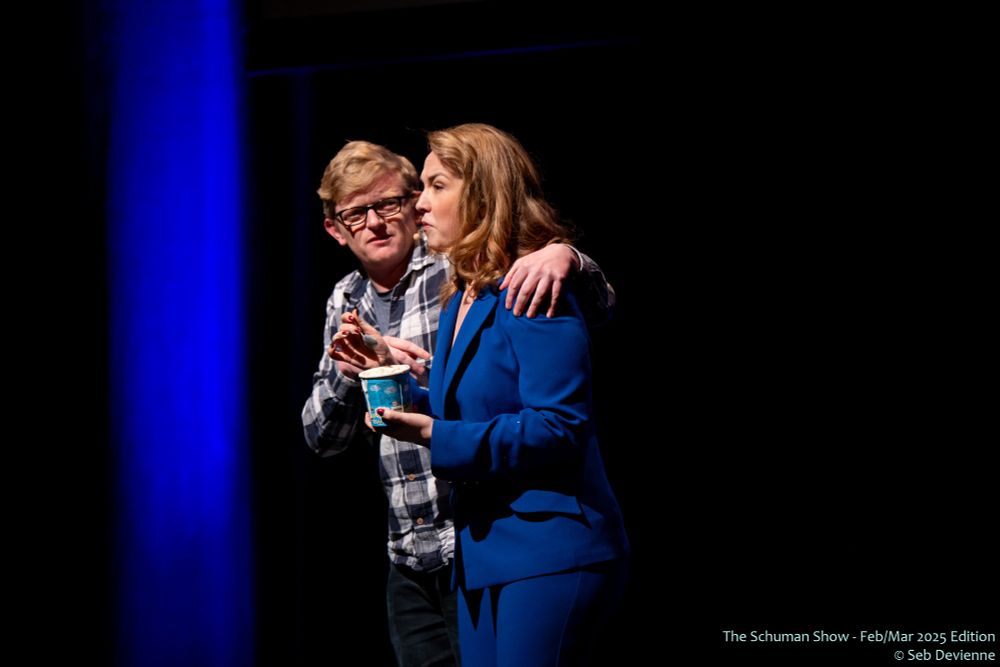 Schuman Show host on stage looking sad, with a fellow cast member feeding her ice cream with his comforting hand on her shoulder